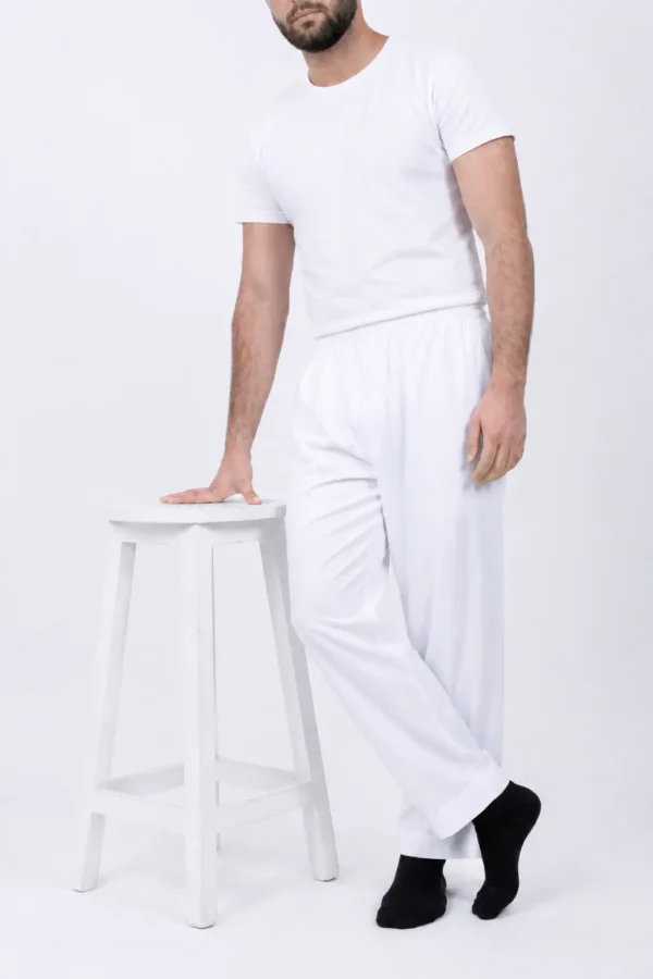Model in an all-white outfit (fitted white t-shirt and wide white pants) standing with one hand resting on a white stool against a light backdrop, wearing black socks.