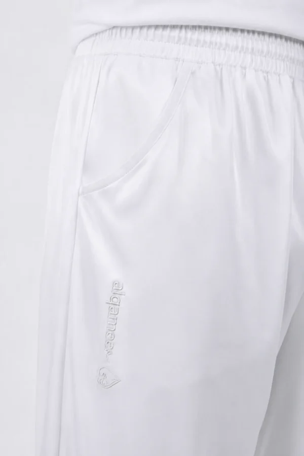 Close-up of white pants showing an elastic waistband and a curved front pocket with a vertical embroidered brand name along the side and a small emblem nearby.