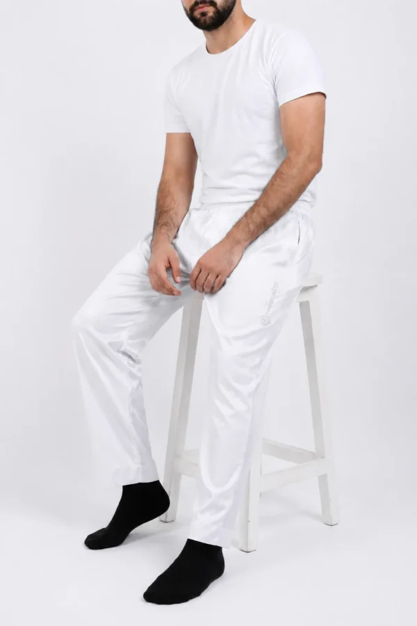 Man in a white T-shirt and white track pants seated on a white stool against a white backdrop, wearing black socks.