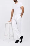 Man wearing all-white loungewear (tee and joggers) leaning with one hand on a white stool, wearing black socks.