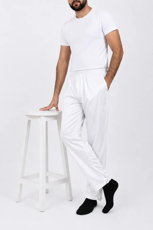 Man wearing all-white loungewear (tee and joggers) leaning with one hand on a white stool, wearing black socks.