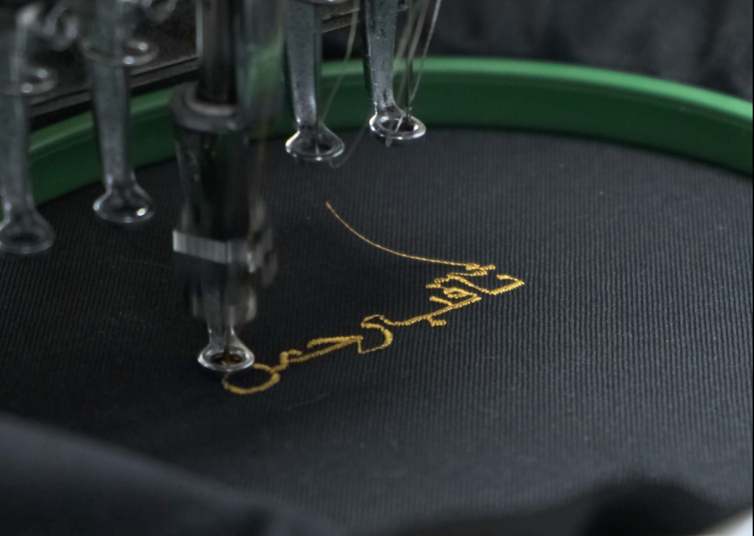 Custom gold Arabic calligraphy embroidery on a black thobe using a precision tailoring machine by alqamees