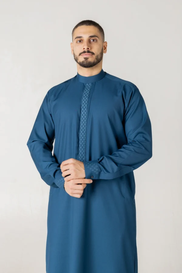 Men’s teal blue thobe with geometric embroidery by alqamees. A ready-to-wear thobe featuring a tailored fit, custom embroidered placket and cuffs, and a classic mandarin collar from the alqamees signature collection.