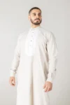 Luxury cream thobe with a white pleated front bib detail by alqamees. A ready-to-wear formal thobe featuring gold buttons, a tailored fit, and a mandarin collar, showcasing the signature craftsmanship of the alqamees collection.