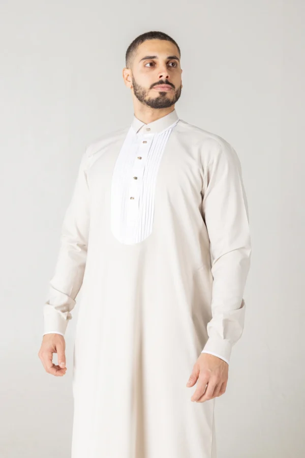 Luxury cream thobe with a white pleated front bib detail by alqamees. A ready-to-wear formal thobe featuring gold buttons, a tailored fit, and a mandarin collar, showcasing the signature craftsmanship of the alqamees collection.