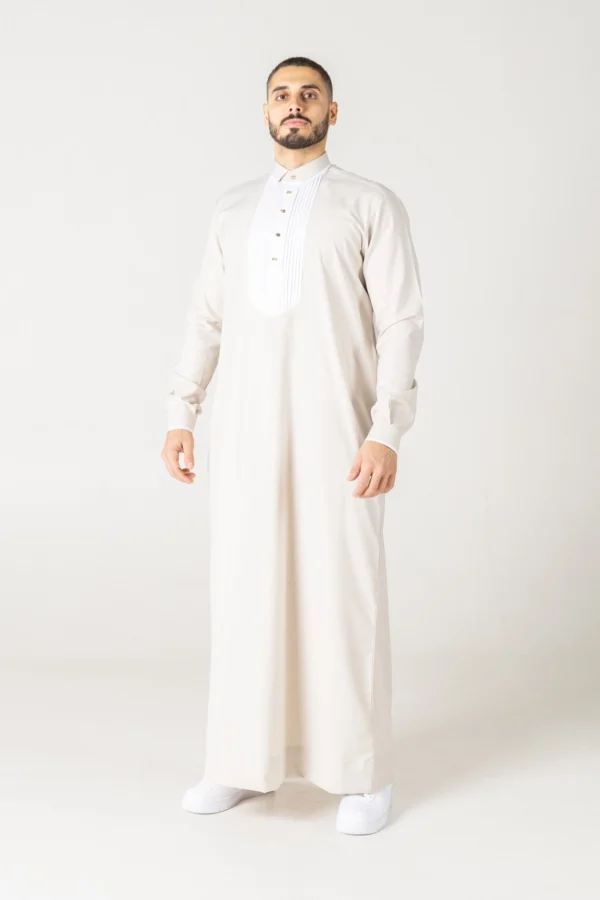 Luxury cream thobe with a white pleated front bib detail by alqamees. A ready-to-wear formal thobe featuring gold buttons, a tailored fit, and a mandarin collar, showcasing the signature craftsmanship of the alqamees collection.