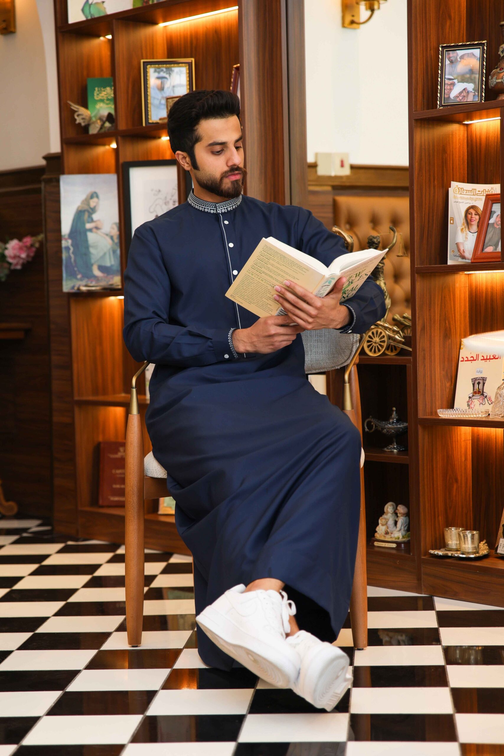 A man with a beard in a navy traditional outfit sits on a chair, reading a book in a wood-paneled room with framed photos on shelves and a black-and-white checkered floor.