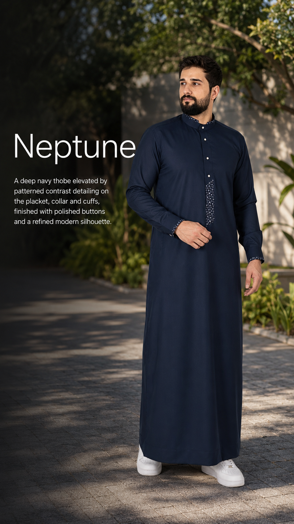 Man in a navy, ankle-length tunic with patterned placket and cuffs stands outdoors among trees.