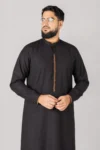 Man wearing a black kurta with orange embroidered placket, glasses, and a beard, standing against a gray backdrop.