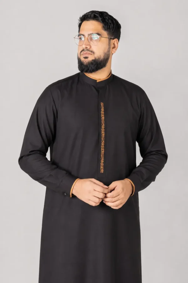 Man wearing a black kurta with orange embroidered placket, glasses, and a beard, standing against a gray backdrop.