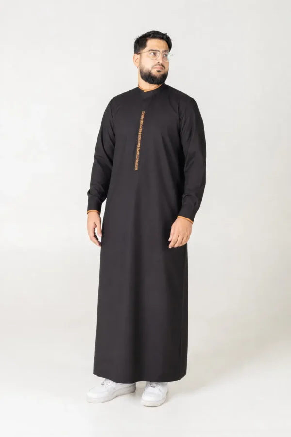 Man wearing a black traditional kandura with orange trim, standing against a light backdrop.