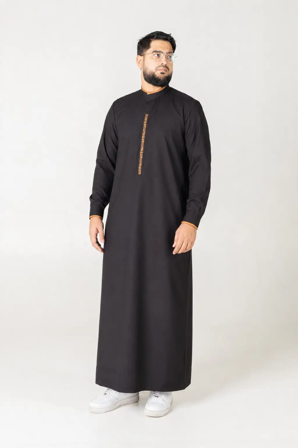 Hawk Black (2) Man wearing a black traditional kandura with orange trim, standing against a light backdrop.