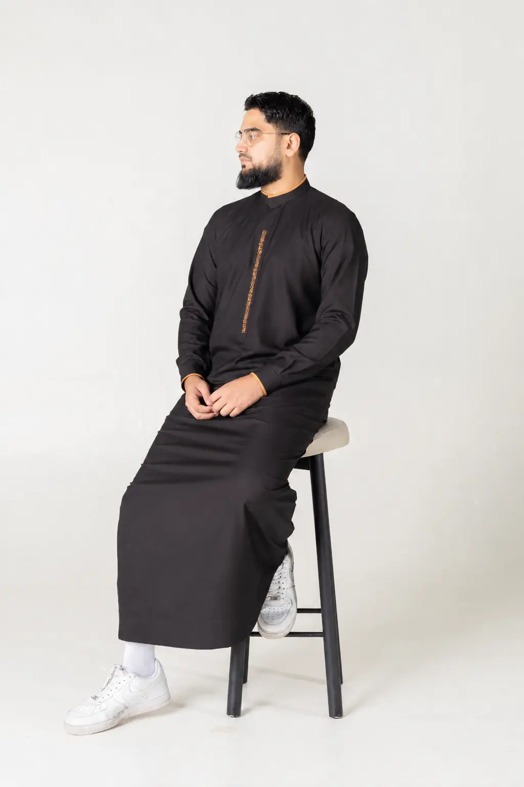 Hawk Black (6) Man in a black kurta with orange embroidery sits on a stool, facing left, wearing white sneakers in a studio with a light background.
