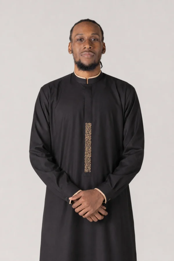Portrait of a man in a black thobe with beige trim and gold vertical embroidery, standing with hands clasped.