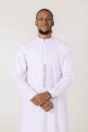 Portrait of a man in a light lavender thobe standing with hands clasped, neutral background behind him.