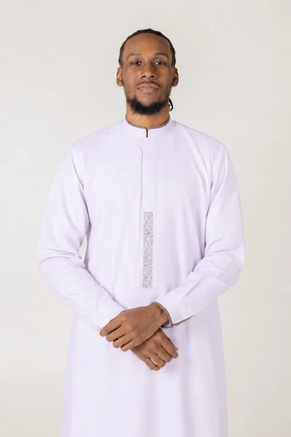 Portrait of a man in a light lavender thobe standing with hands clasped, neutral background behind him.