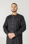 Man wearing a black thobe with a tan collar stands against a light gray background, hands loosely clasped.