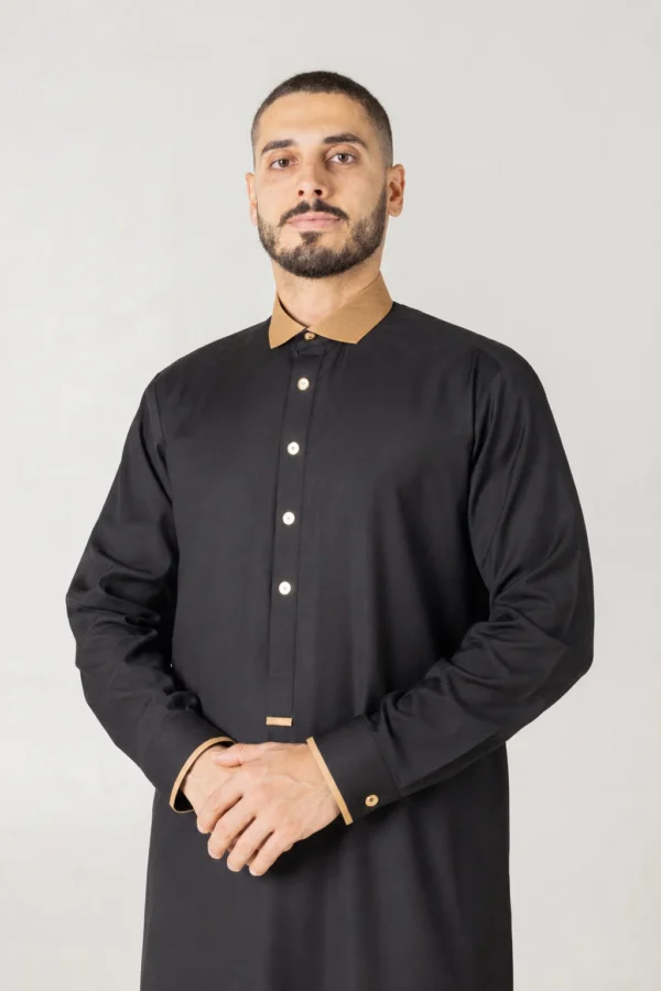 Man wearing a black thobe with a tan collar stands against a light gray background, hands loosely clasped.