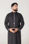 Man in a black traditional kurta with white geometric embroidery on the placket and cuffs, standing with hands clasped, looking to the right.