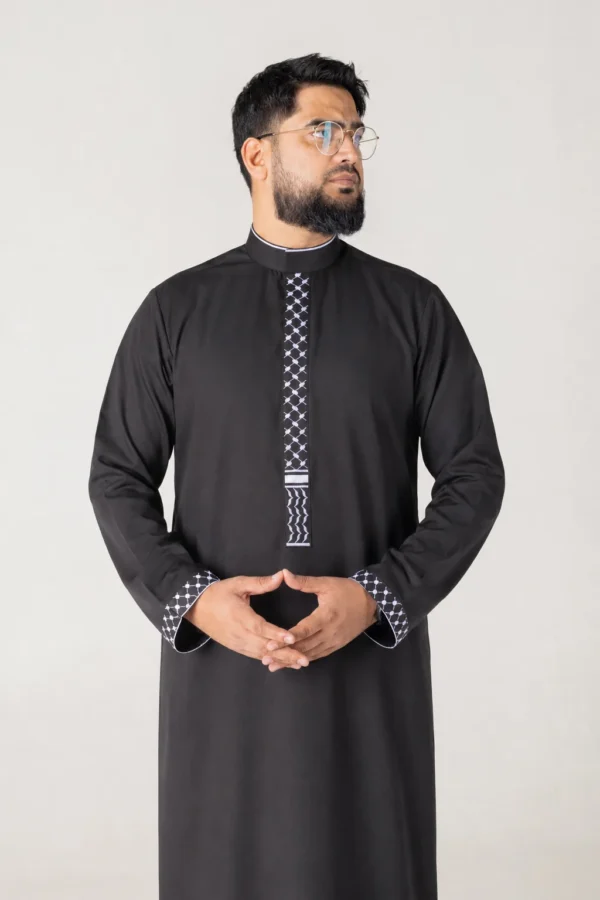 Man in a black traditional kurta with white geometric embroidery on the placket and cuffs, standing with hands clasped, looking to the right.
