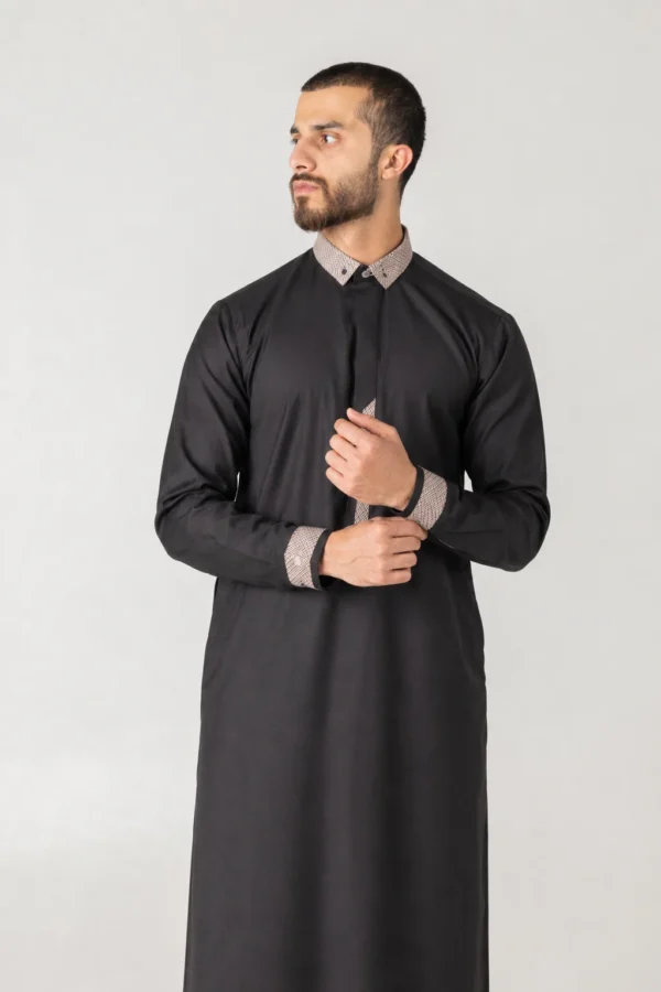 Man wearing a black thobe with beige patterned collar and cuffs, standing against a light background and looking to his left.