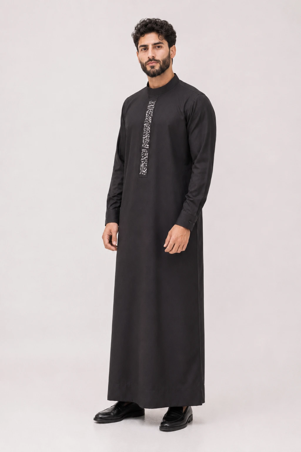 Backhour Black (3) Man standing in a black thobe with a vertical white embroidered panel on the chest, wearing black shoes.