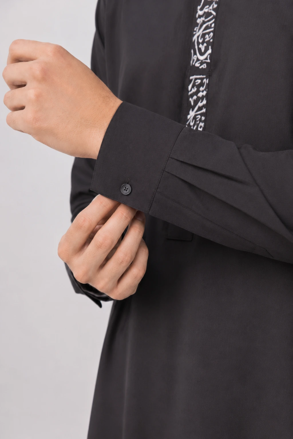 Backhour Black (5) Close-up of a person buttoning the cuff of a black garment with white embroidered trim.
