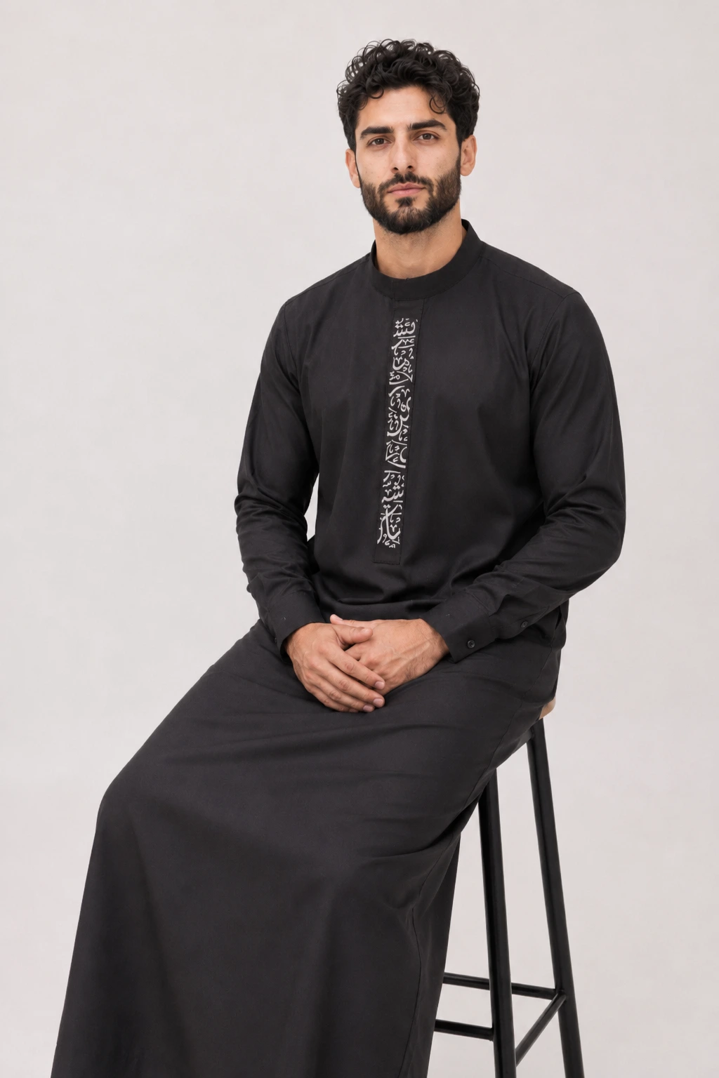 Backhour Black (6) Man in a black traditional robe with white vertical embroidery, seated on a stool against a light backdrop.