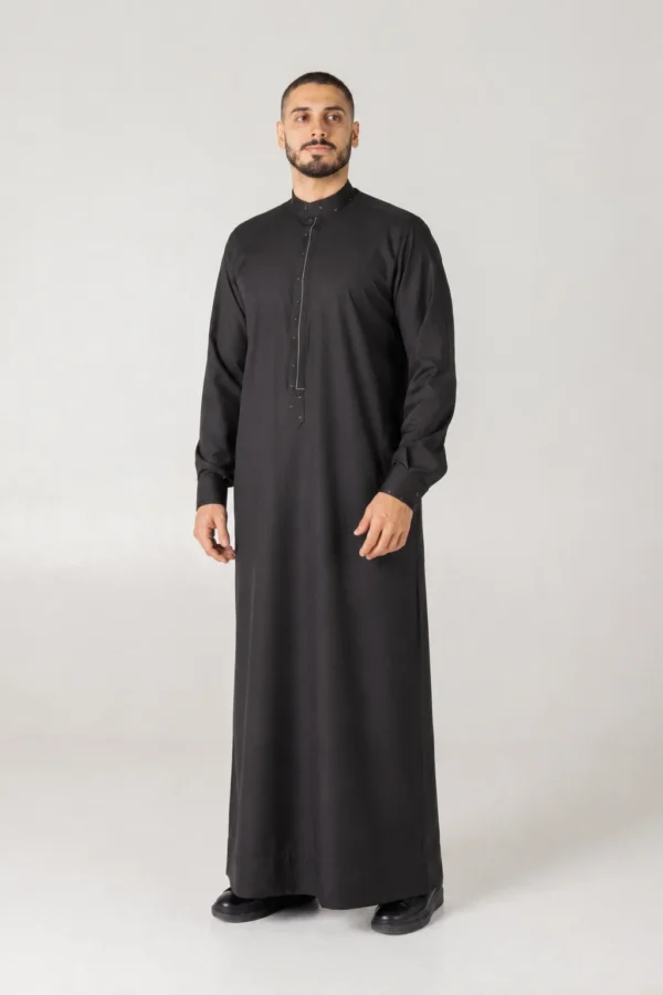 Full-length view of a man in a black thobe standing against a pale gray backdrop.