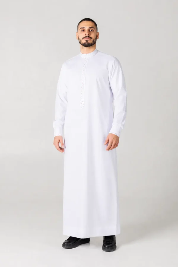 Man wearing a white thobe with decorative embroidery on the collar and cuffs, standing against a light gray background.