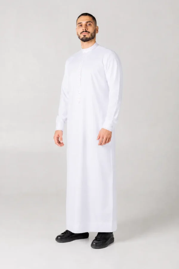 Full-length image of a man wearing a white thobe with embroidered collar against a light gray background, standing with hands at his sides, black shoes visible.
