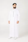 Bearded man in a white thobe with a vertical decorative stripe, standing against a light background, looking to the side.