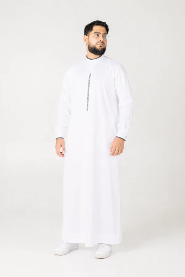 Bearded man in a white thobe with a vertical decorative stripe, standing against a light background, looking to the side.