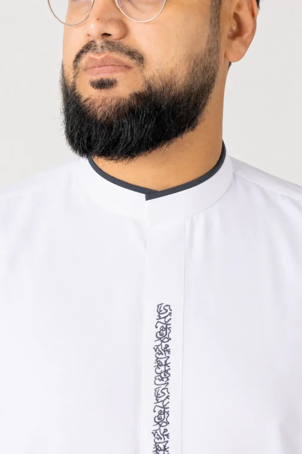 Close-up of a man wearing a white thobe with black collar trim and decorative embroidered front placket; glasses are visible at the top edge of the image.