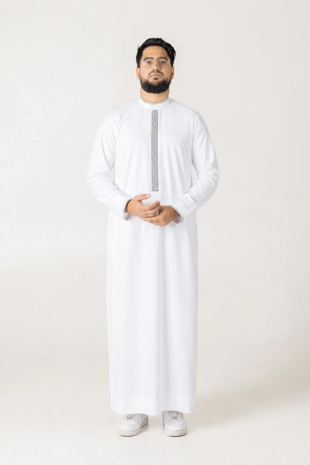 Nexus white 1 Man standing in a plain studio wearing a white thobe with decorative vertical embroidery, hands lightly clasped in front, wearing white sneakers.