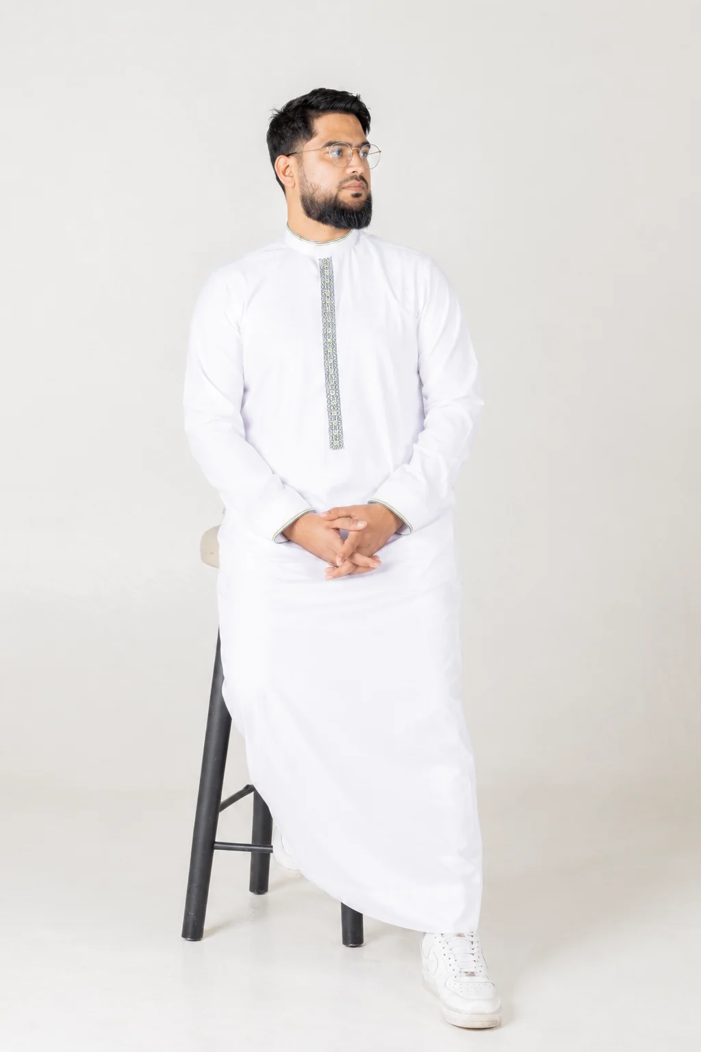 Nexus white 5 Man in a white kurta with embroidered placket sits on a black stool, hands clasped, looking to the left.