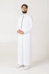 Man standing in a white thobe with a decorative vertical embroidery stripe, wearing glasses and with hands loosely clasped in front, neutral background.