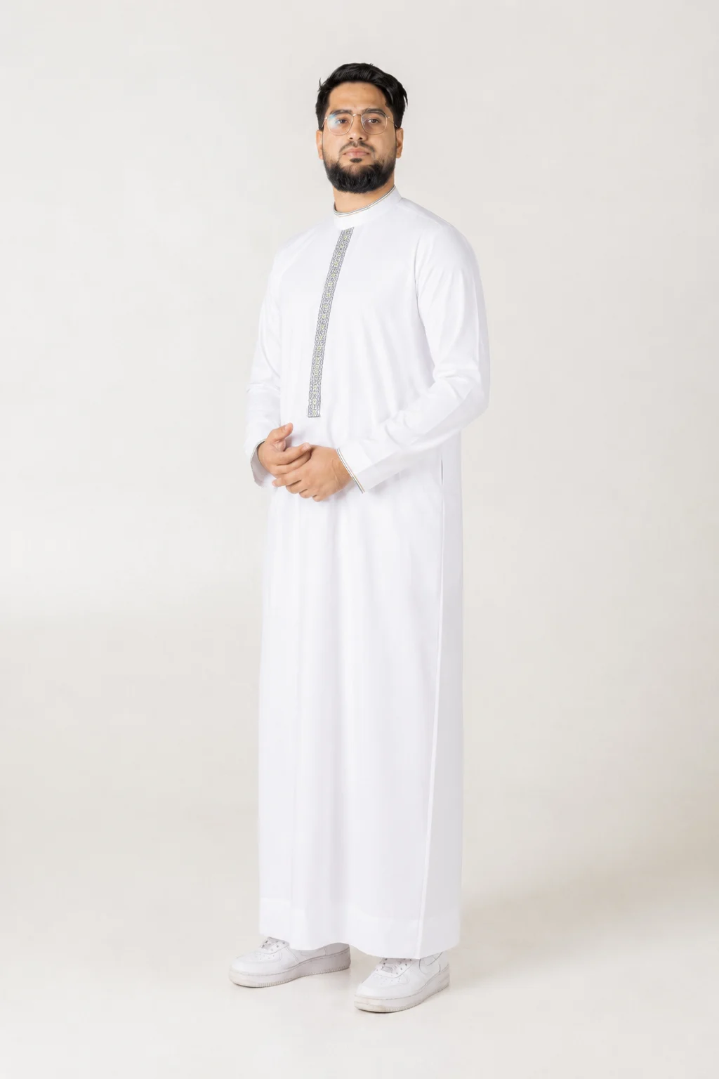 Nexus white 6 Man standing in a white thobe with a decorative vertical embroidery stripe, wearing glasses and with hands loosely clasped in front, neutral background.