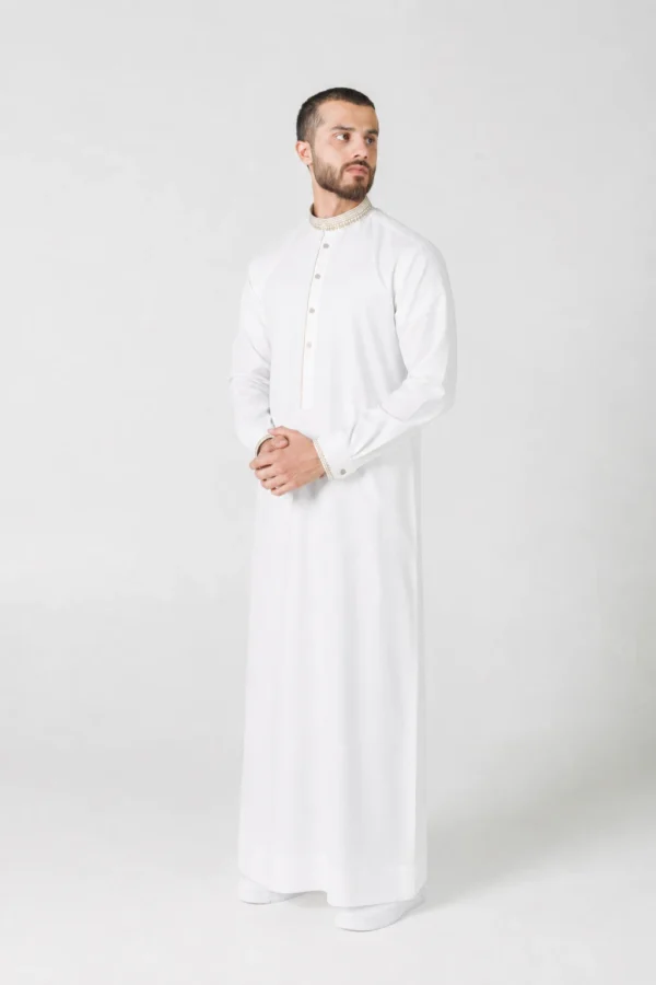 Man in a full-length white thobe with beige embroidered collar and cuffs, standing with hands clasped.