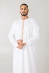 Man in a white kandura with beige embroidered trim down the front and on the cuffs, hands clasped in front.