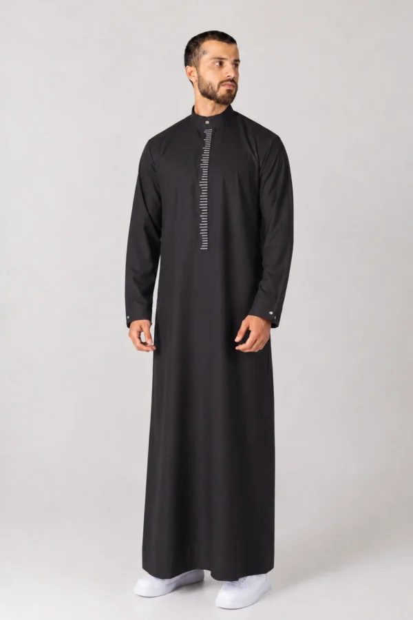 Man wearing a black abaya-style thobe with a vertical white striped placket, standing against a light gray backdrop.
