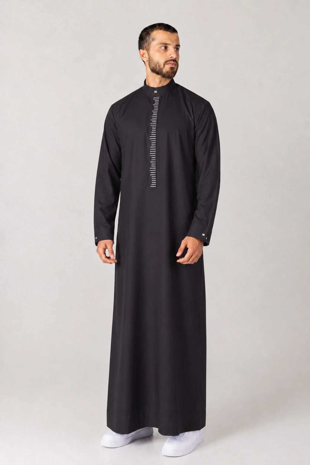 Rasasi Black Edition (3) Man wearing a black abaya-style thobe with a vertical white striped placket, standing against a light gray backdrop.