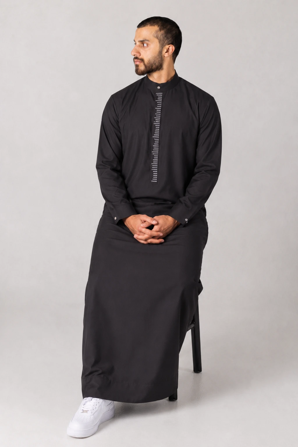Rasasi Black Edition (7) Man in a black kurta seated on a chair, looking to his left, with a vertical white measuring-scale design on the chest and white sneakers visible at the feet.