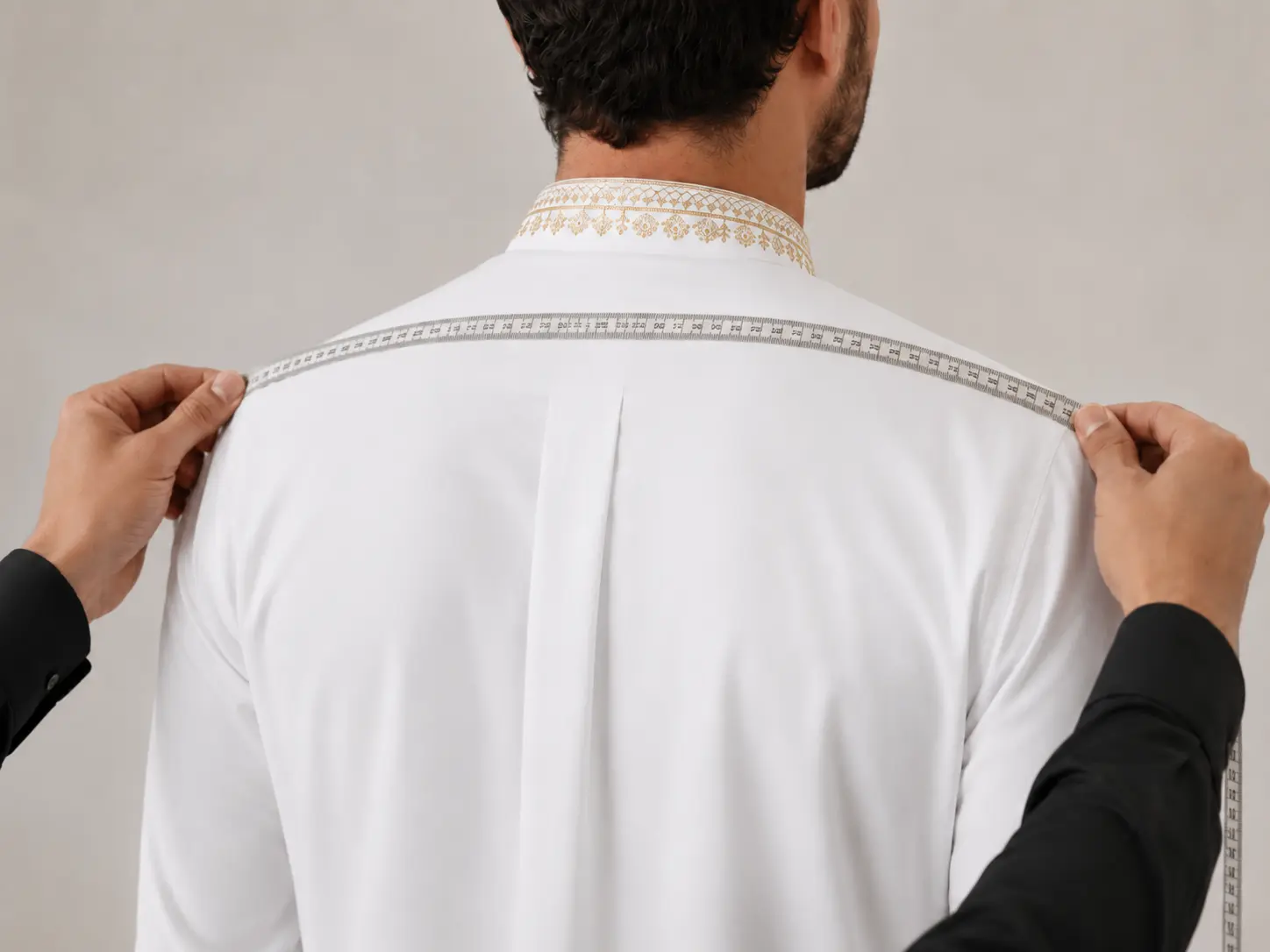 Tailor measures a man's shoulders with a tape measure across a white shirt from behind, assessing fit.